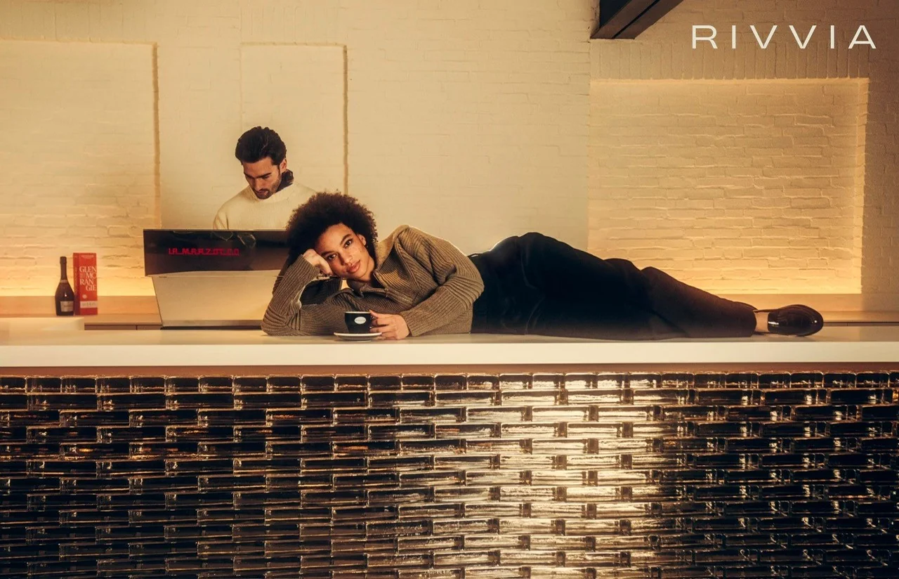 Vrouw ligt met een kopje koffie op een moderne bar terwijl een barista werkt achter een La Marzocco koffiemachine in een stijlvolle ruimte met het logo van RIVVIA op de muur.