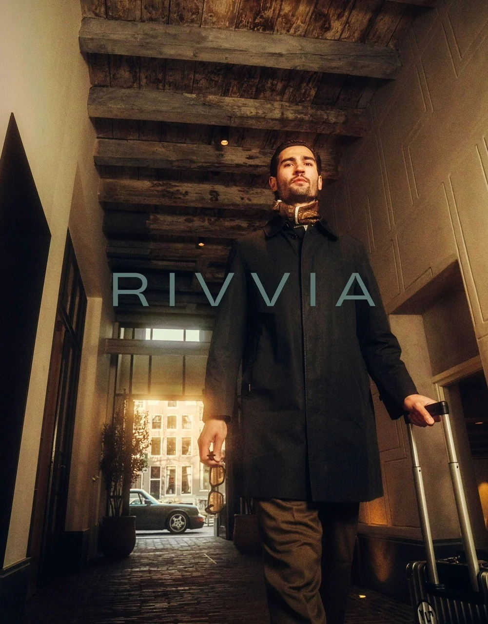 Man in stijlvolle outfit trekt een koffer achter zich aan in een sfeervolle stadse entree met het logo "RIVVIA" op de voorgrond.