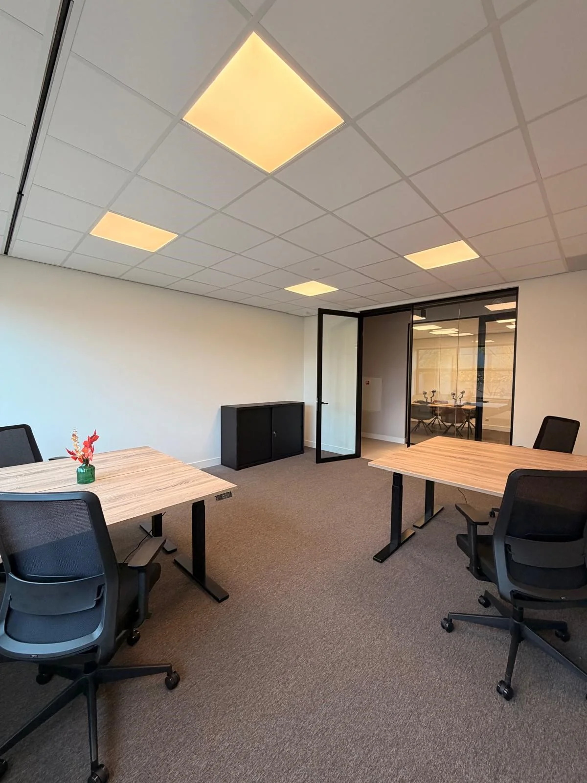 Modern office space with four desks, ergonomic chairs, and a glass door leading to a conference room.