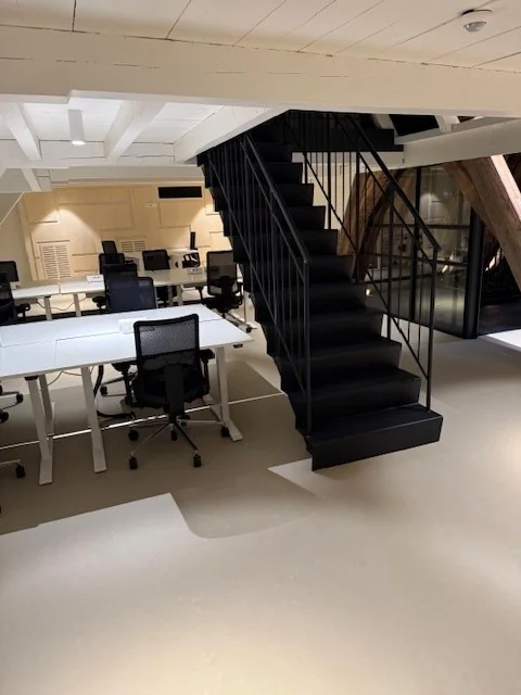Moderne kantoorruimte met witte bureaus, zwarte bureaustoelen en een zwarte stalen trap in het midden.