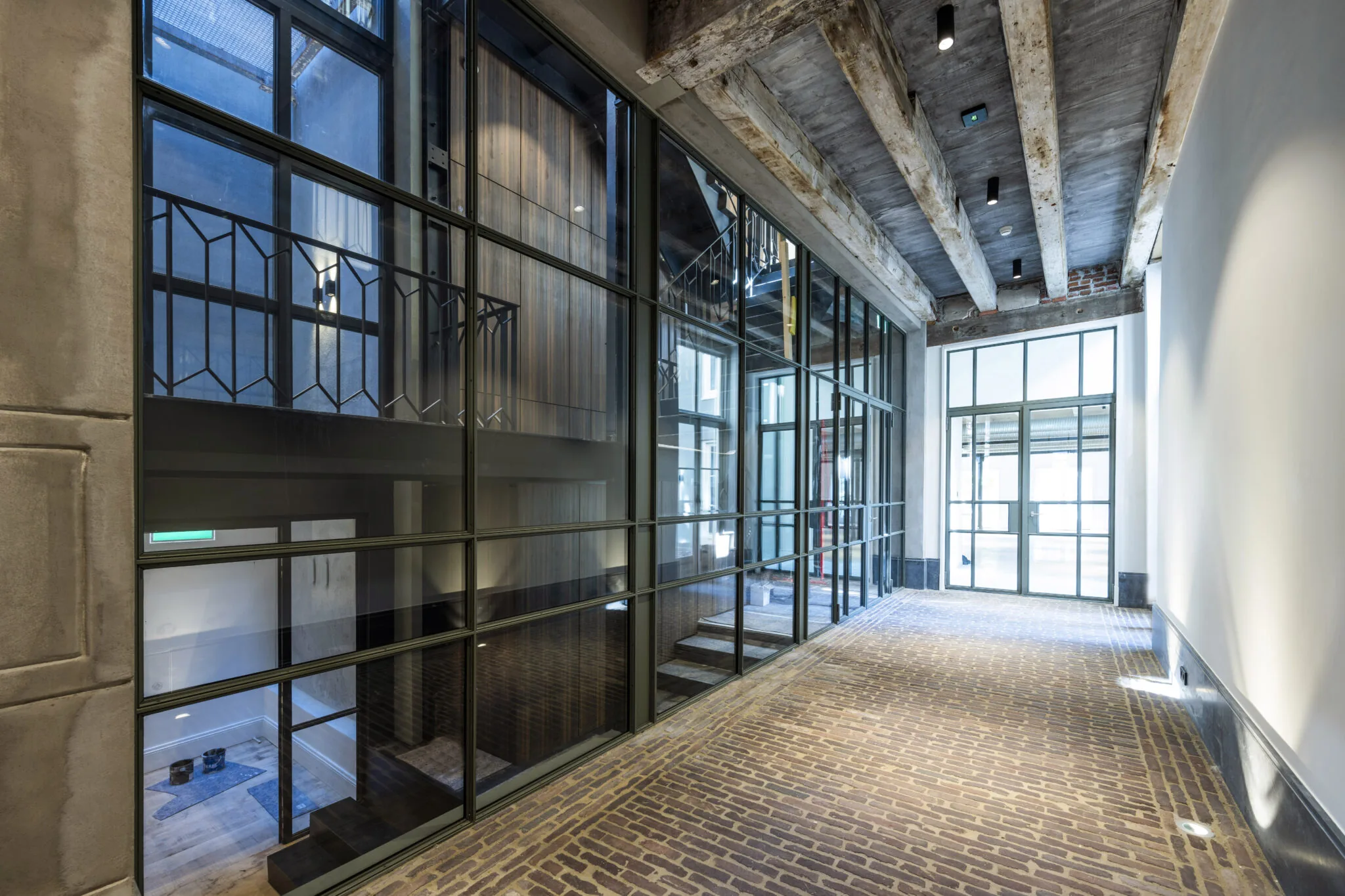 Lichtovergoten gang met bakstenen vloer, glazen wanden en een plafond met houten balken.