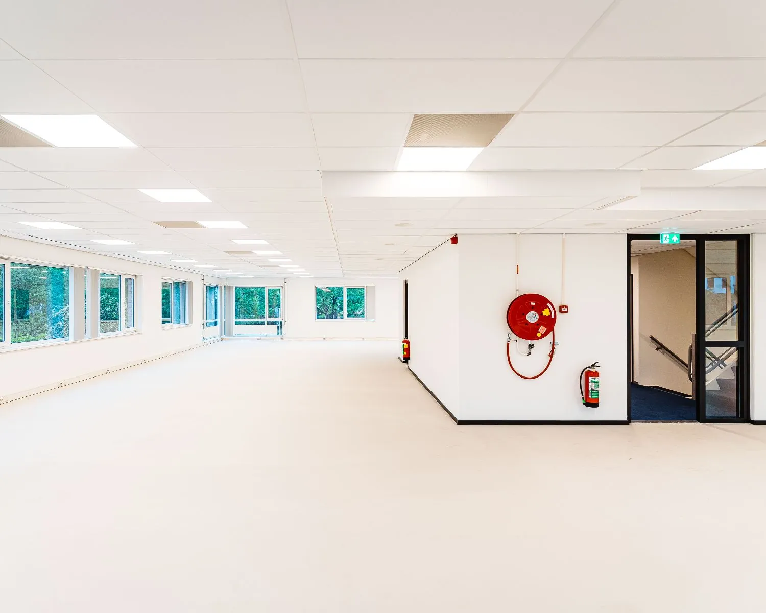 Licht en ruim leeg kantoorpand met grote ramen, systeemplafond en brandhaspel aan de muur.