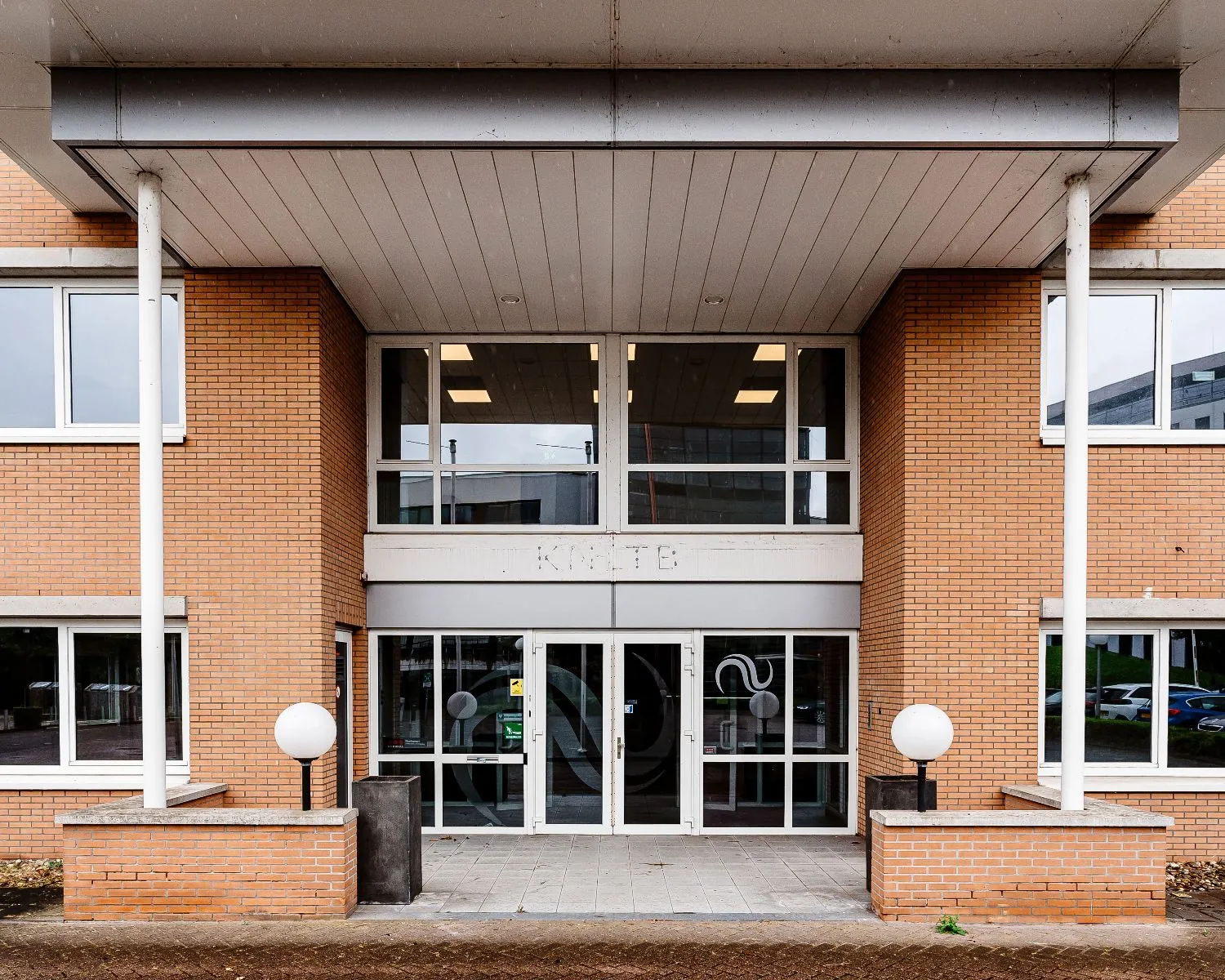 Ingang van een kantoorpand met bakstenen gevel, grote glazen deuren en de naam 'KHTB' boven de entree.