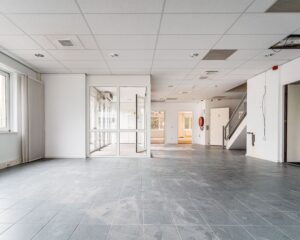 Lichte, lege kantoorruimte met grijze vloertegels, systeemplafond en zicht op een glazen tussenwand en trap.