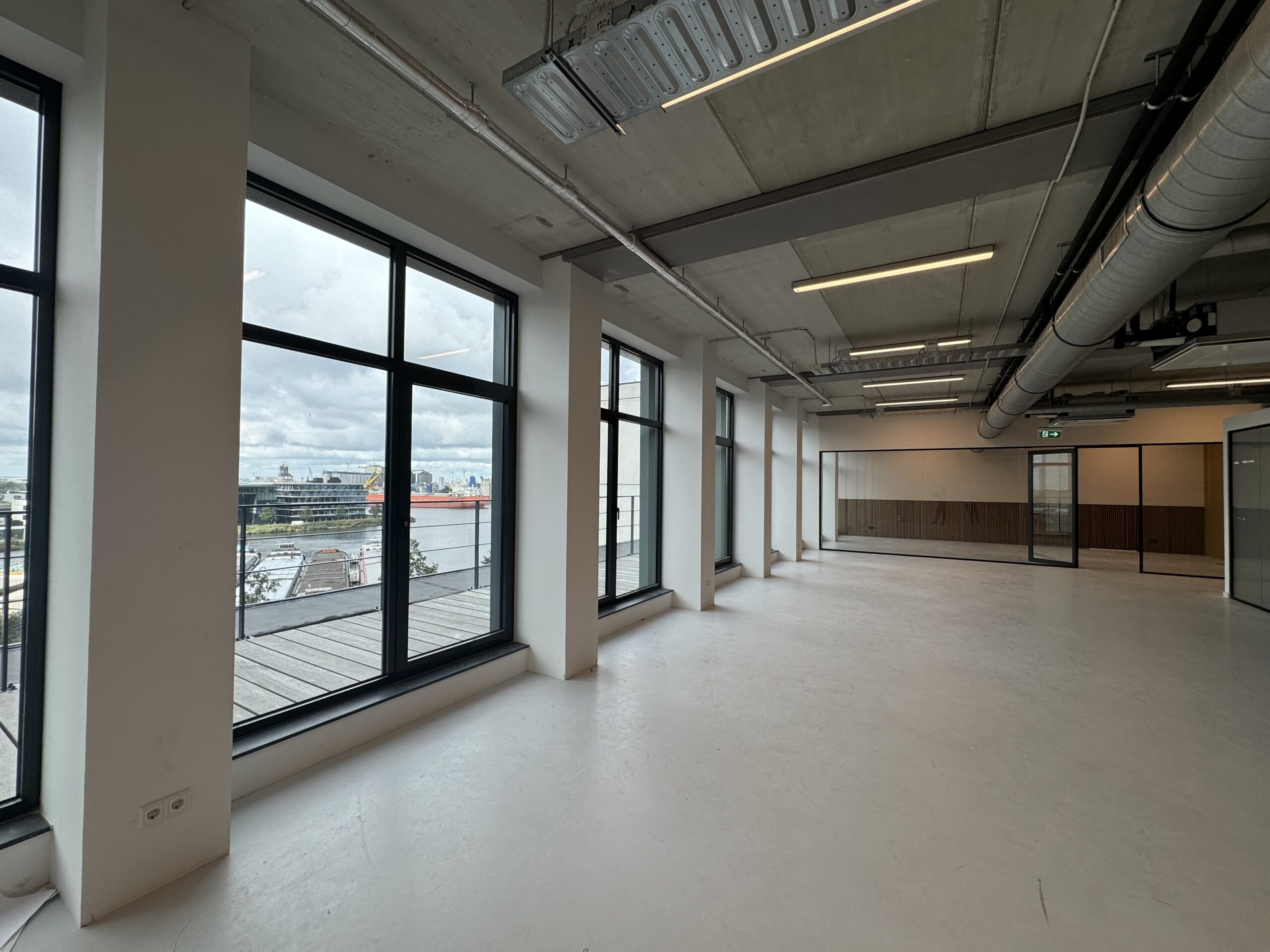 Een lege kantoorruimte met grote ramen en uitzicht op een industrieel gebied en water.