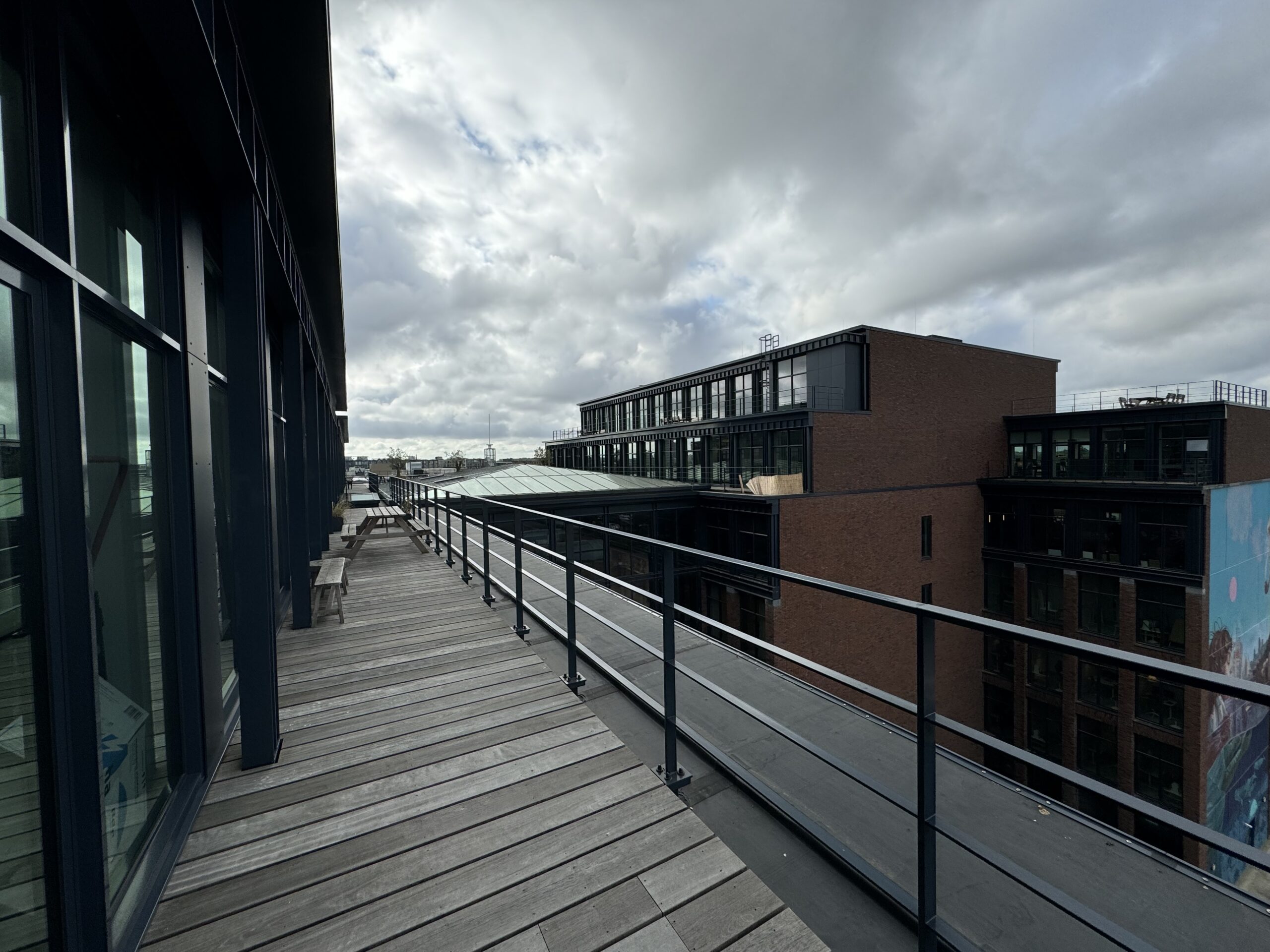 Een houten dakterras met zitbanken naast moderne kantoorgebouwen onder een bewolkte hemel.