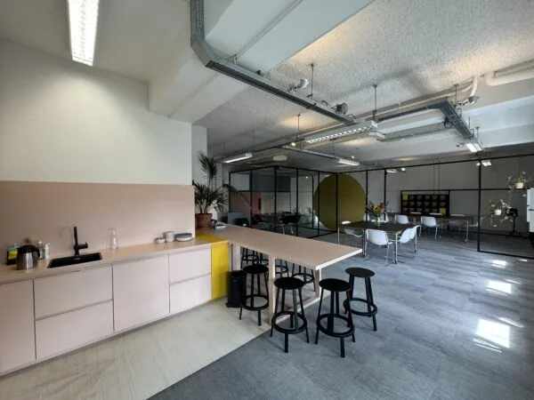 Moderne kantoorruimte met een open keuken, hoge tafel met barkrukken en vergadertafels in een lichte, industriële stijl.