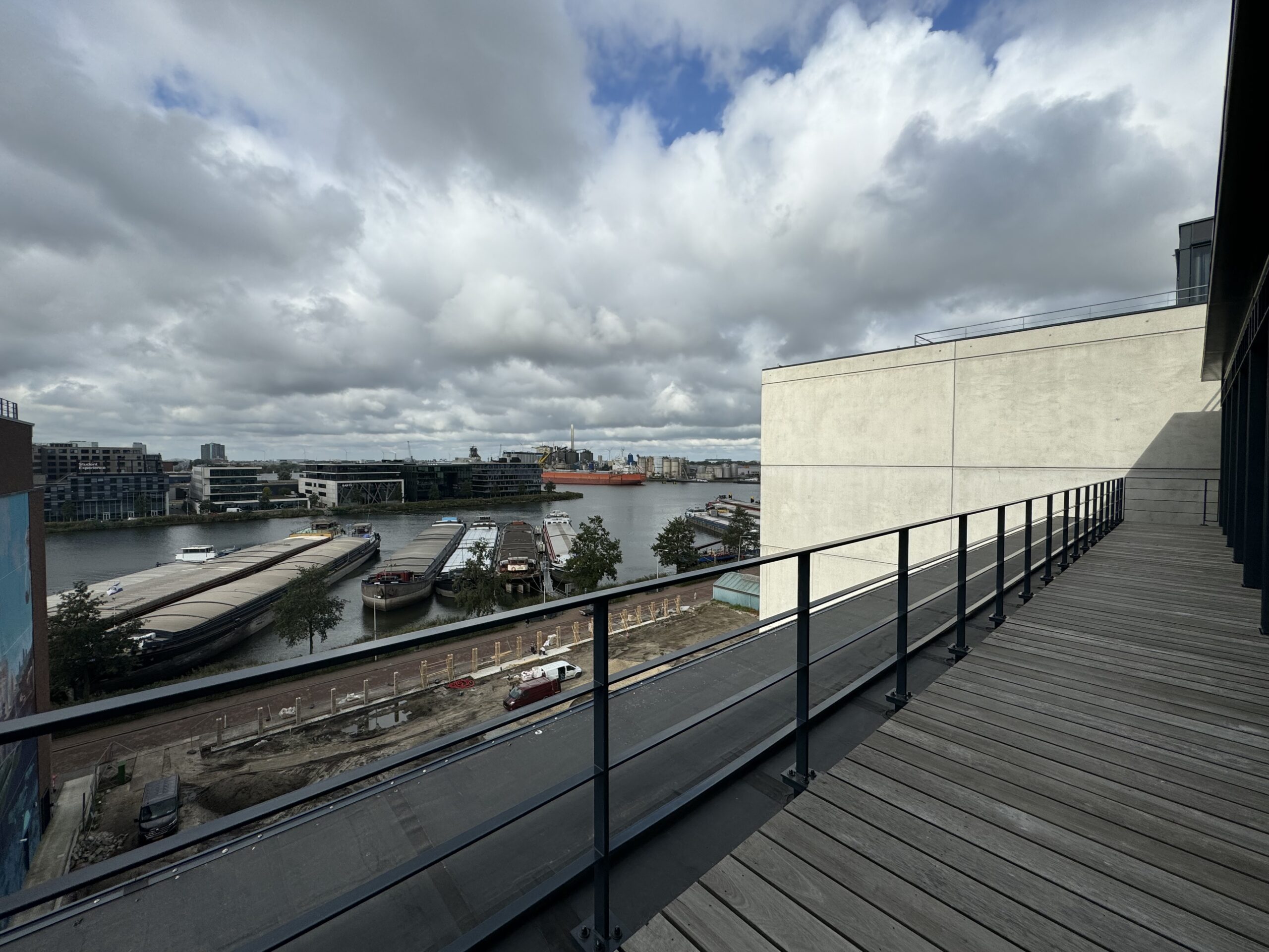 Uitzicht vanaf een houten terras met metalen hek op een rivier met aangemeerde boten onder een bewolkte lucht.