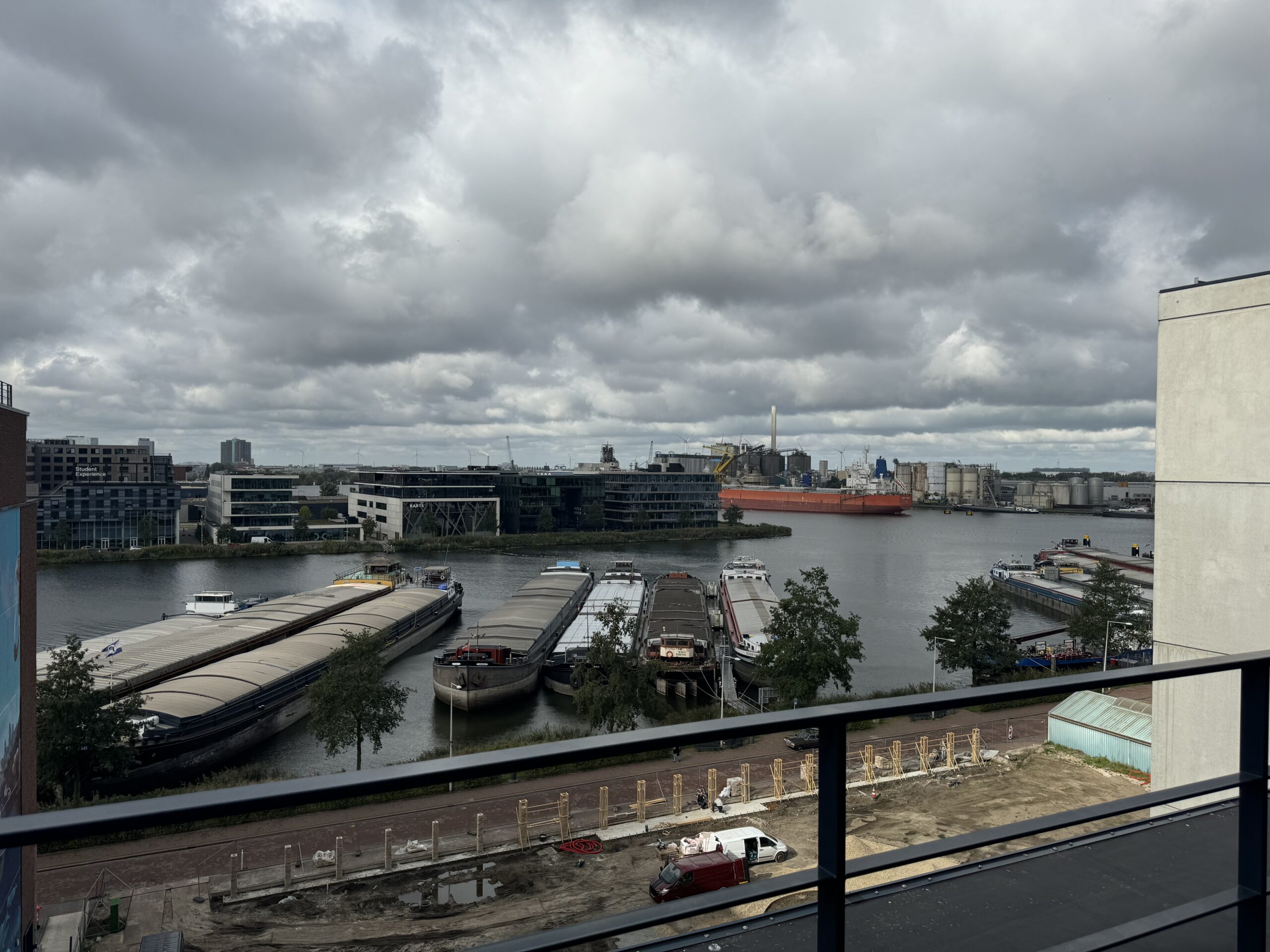 Uitzicht op een rivier met aangemeerde vrachtschepen en industriële gebouwen aan de overkant onder een bewolkte hemel.