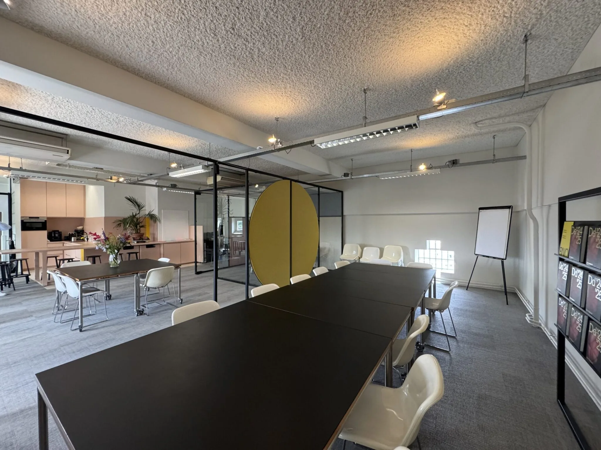 Moderne vergaderruimte met lange zwarte tafel, witte stoelen, flip-over en een open keuken op de achtergrond.