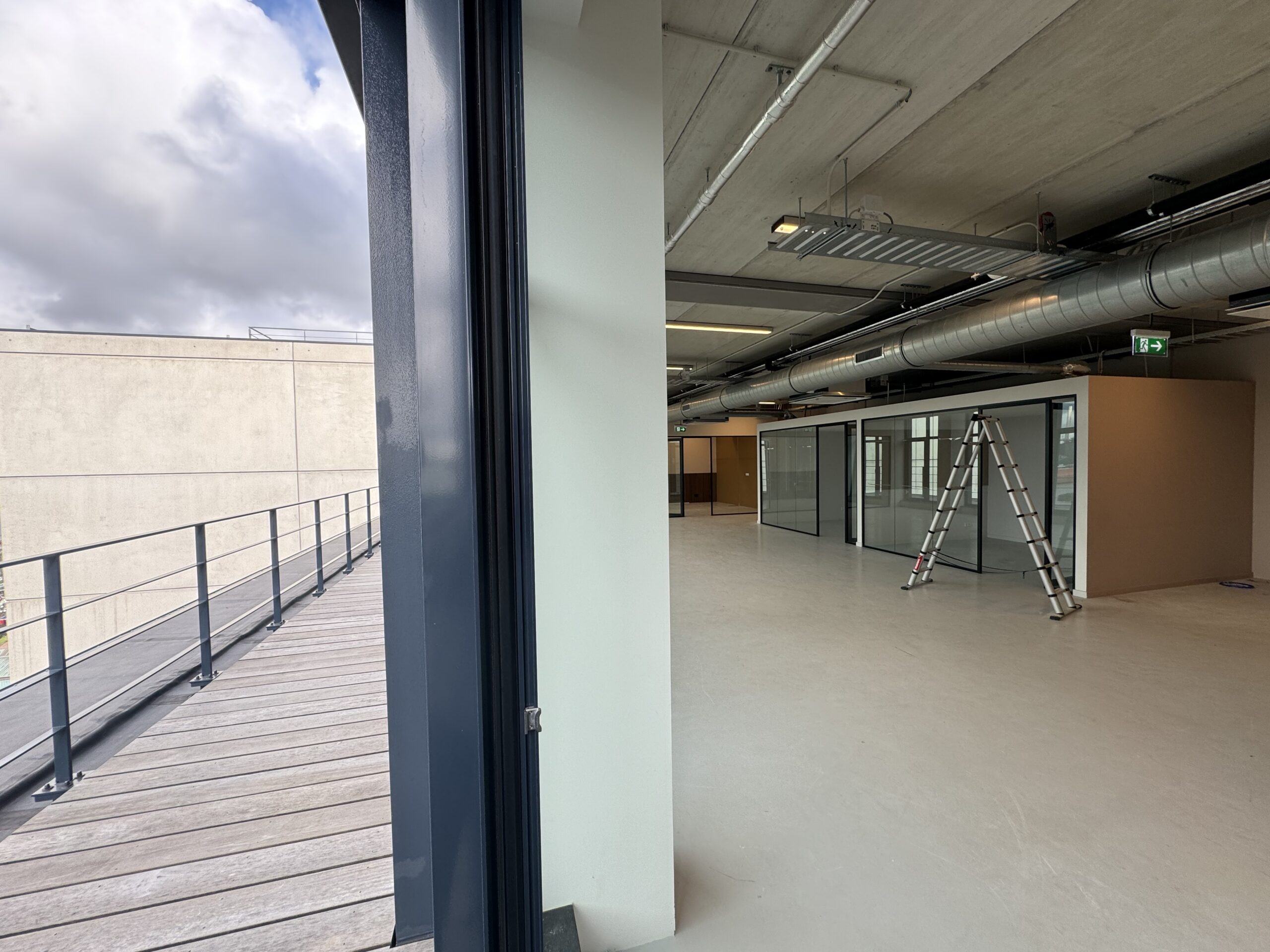 Een leeg kantoorinterieur met glazen wanden en een ladder naast een buitenbalkon onder een bewolkte lucht.