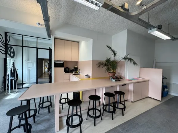 Moderne kantoorkeuken met lichtroze keukenkasten, zwarte barkrukken en een lange tafel in een open ruimte.