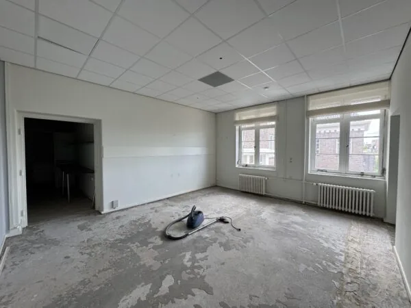 Lege, deels gestripte kamer met een stofzuiger op de vloer en twee grote ramen aan de rechterzijde.