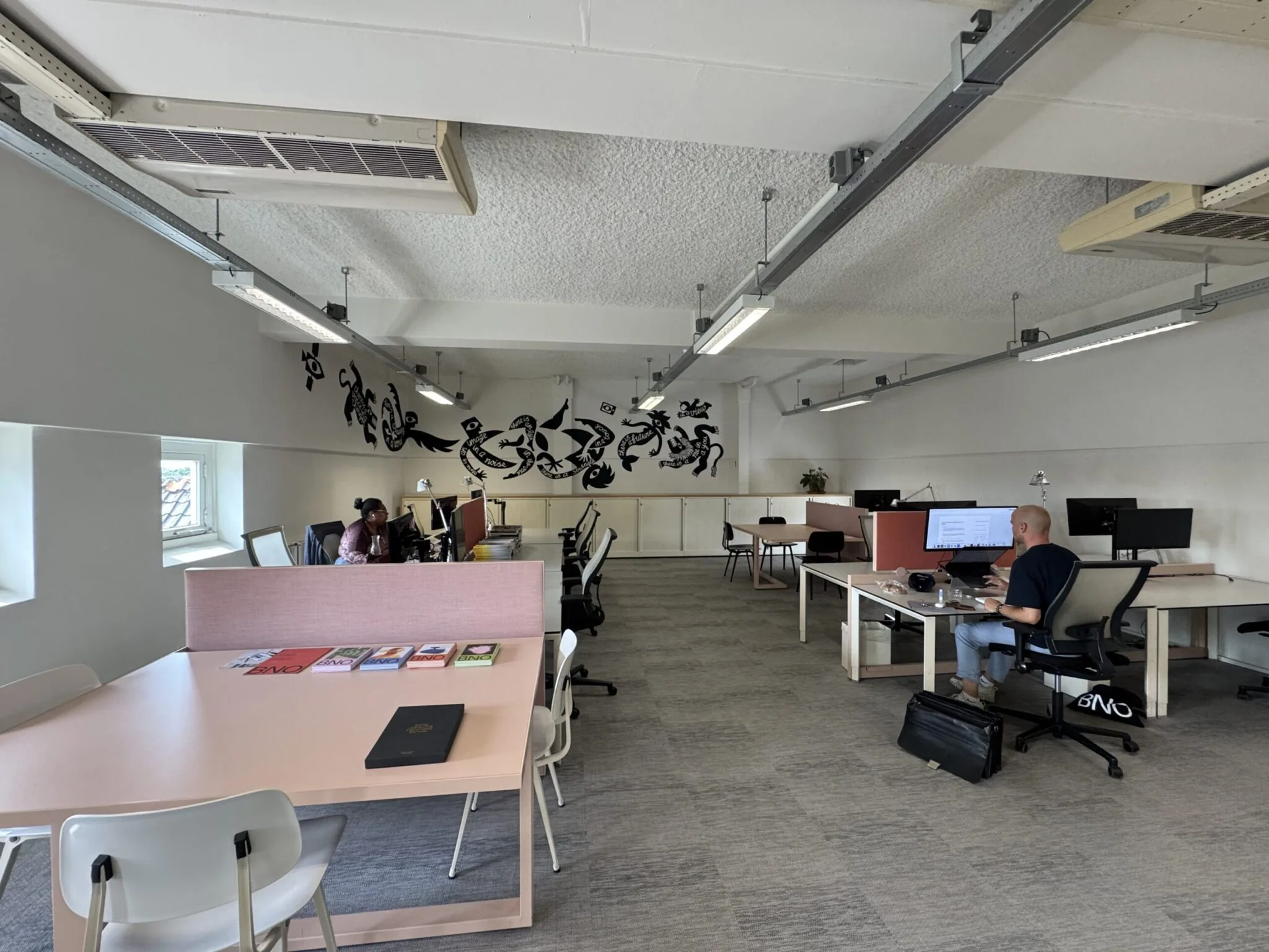 Ruime en moderne kantoorruimte met werkplekken, een muurschildering en enkele mensen aan het werk.