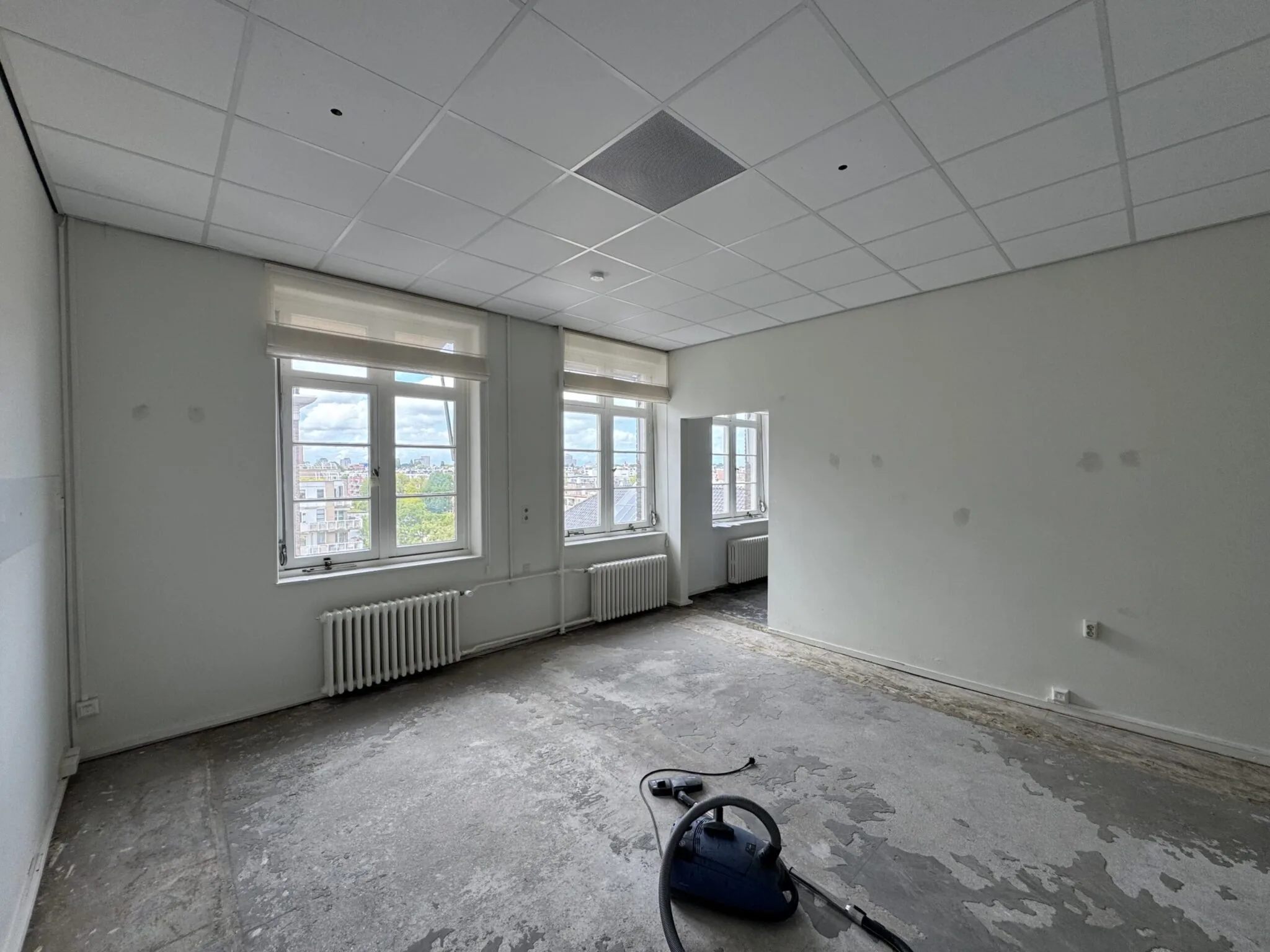Lege, deels gerenoveerde kamer met grote ramen, een kale vloer en een stofzuiger op de grond.