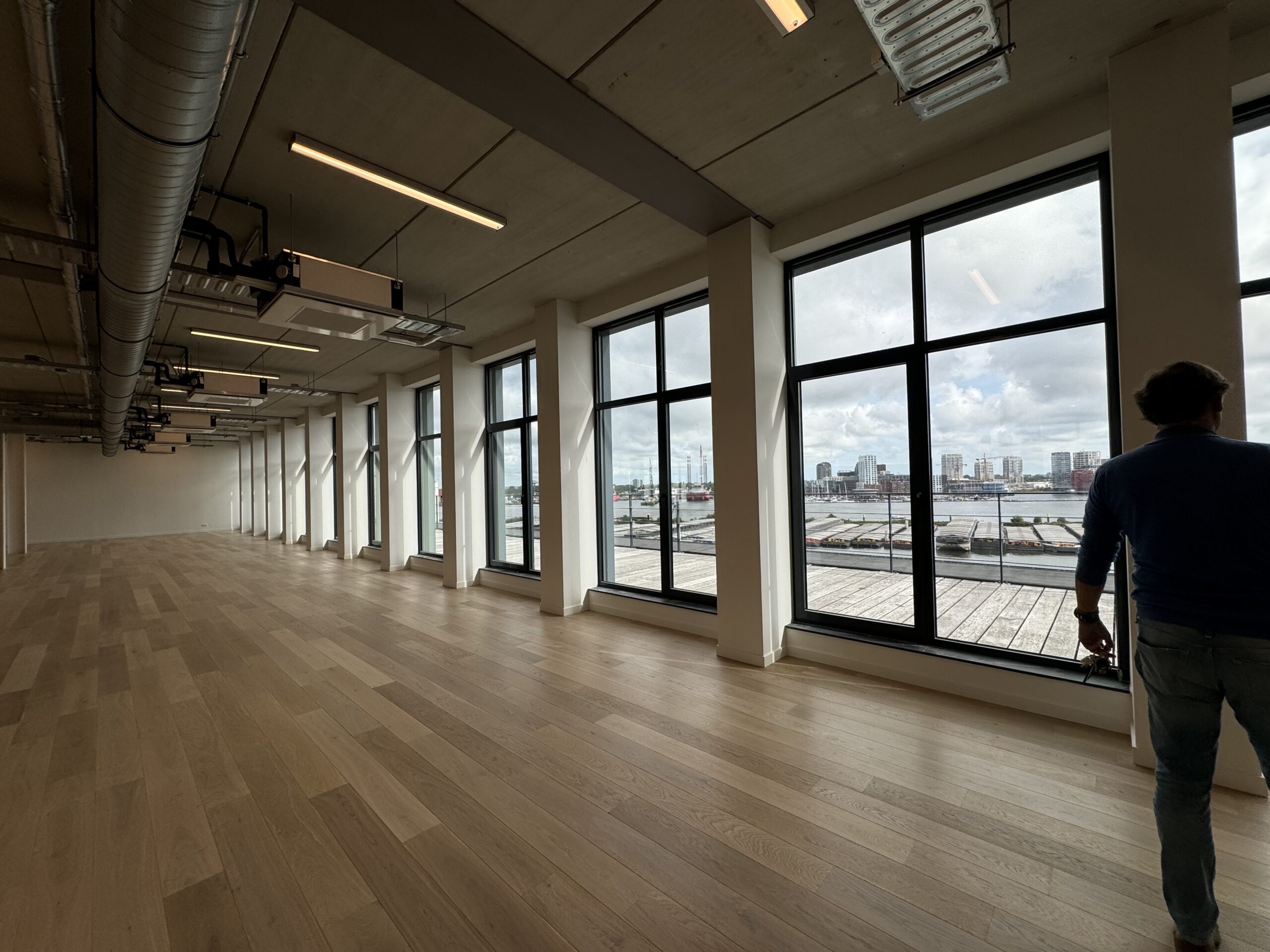 Een leeg kantoor met houten vloer en grote ramen met uitzicht op de stad en de haven.
