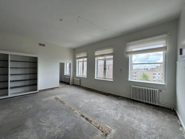 Lege kamer met kale vloer, grote ramen met uitzicht op de stad en een ingebouwde boekenkast.