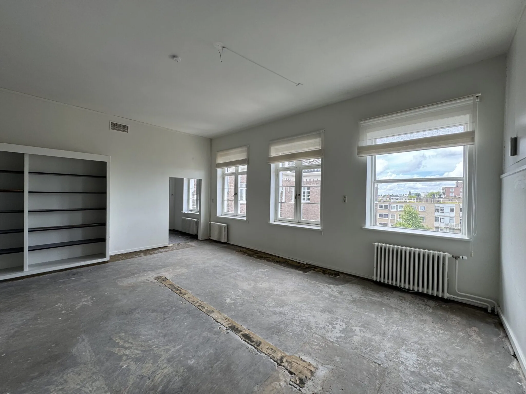 Lege kamer met kale vloer, grote ramen met uitzicht op de stad en een ingebouwde boekenkast.