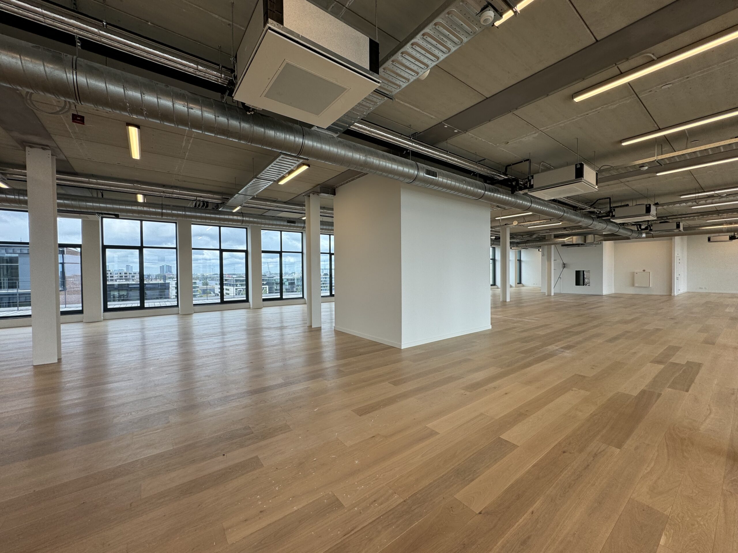 Een ruime, moderne kantoorruimte met houten vloeren en grote ramen met uitzicht op de stad.