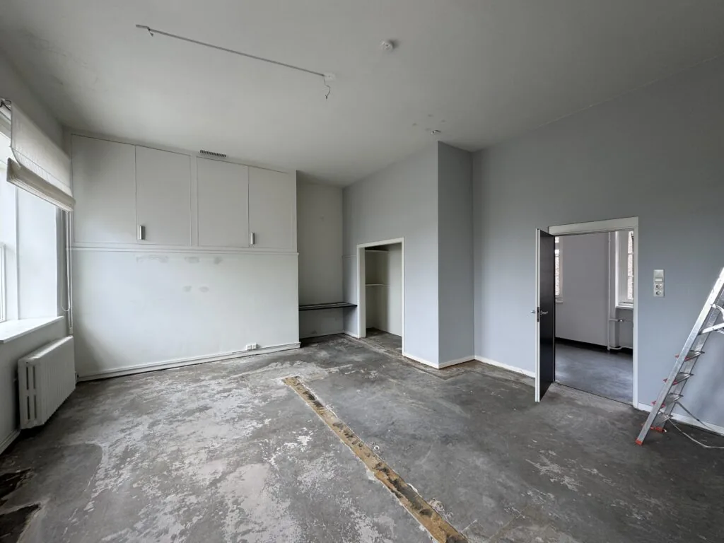 Lege kamer met kale betonvloer, lichtgrijze muren, ingebouwde kast en open deur naar aangrenzende ruimte.