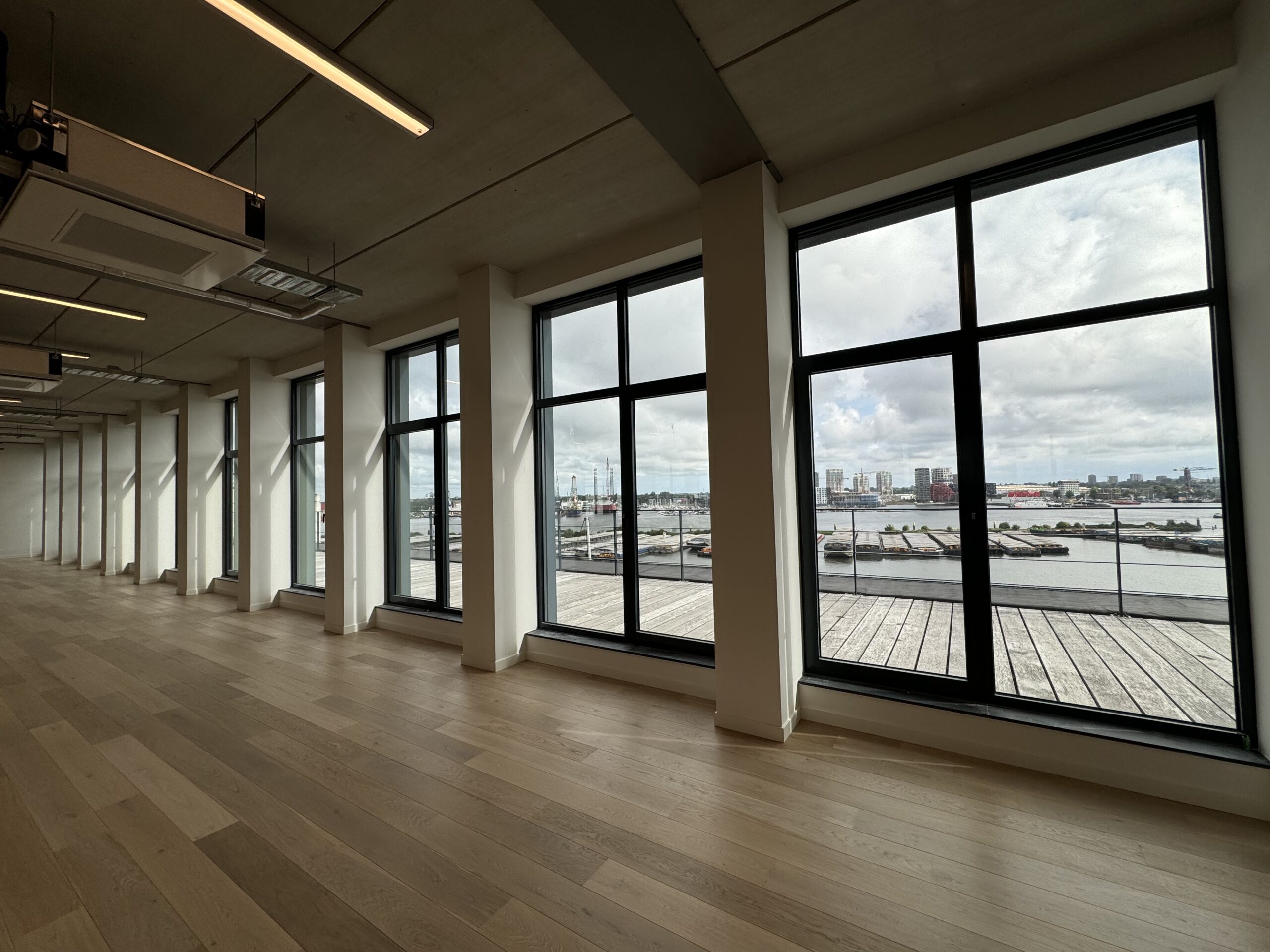 Grote ramen in een ruime kamer bieden uitzicht op een haven en stad aan de horizon.