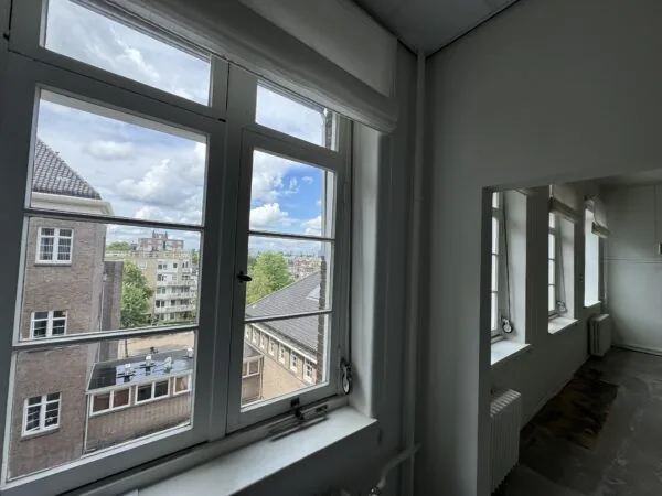 Uitzicht vanuit een lichte kamer met meerdere ramen op een stedelijk landschap met gebouwen en bomen.
