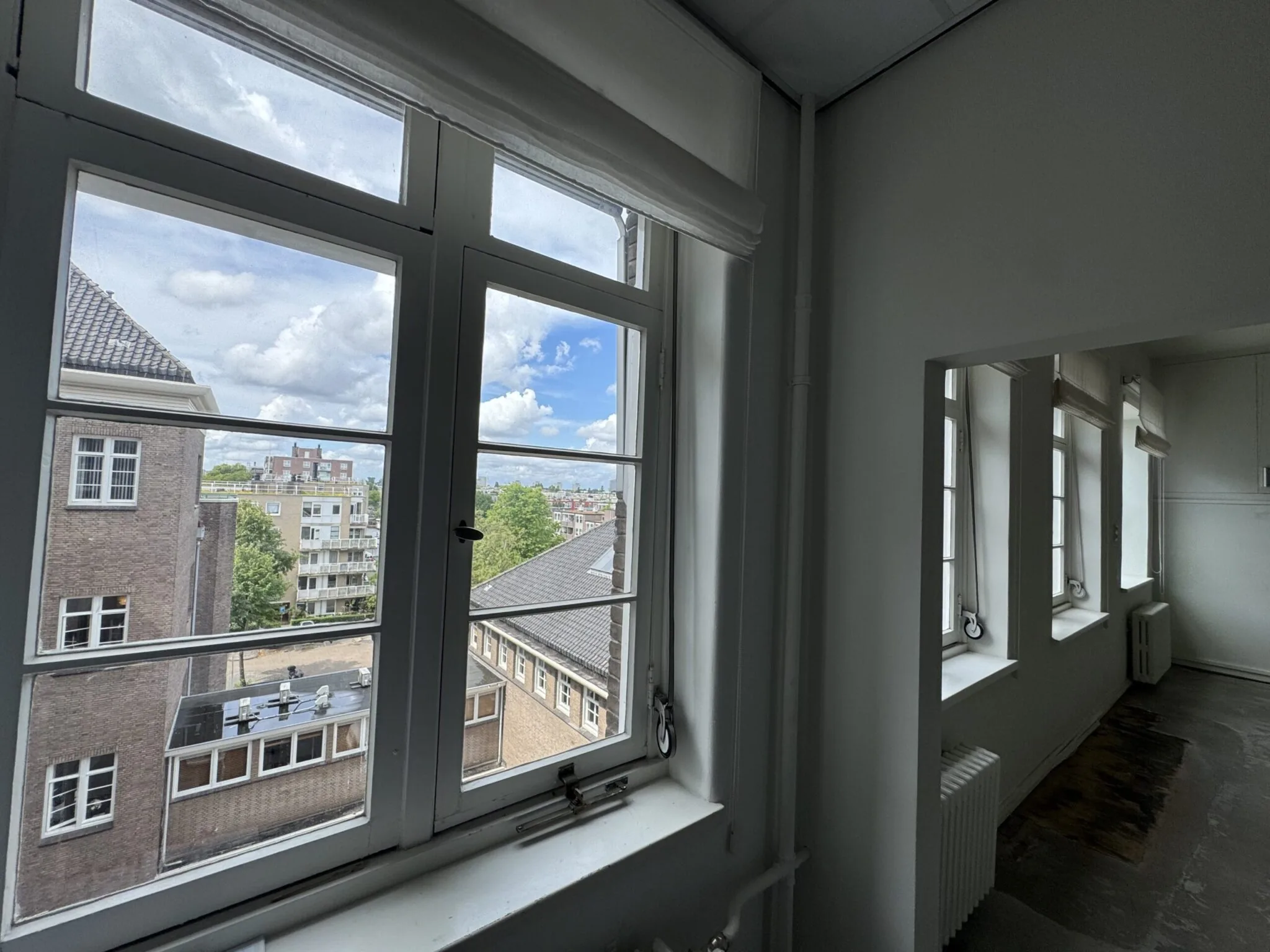 Uitzicht vanuit een lichte kamer met meerdere ramen op een stedelijk landschap met gebouwen en bomen.