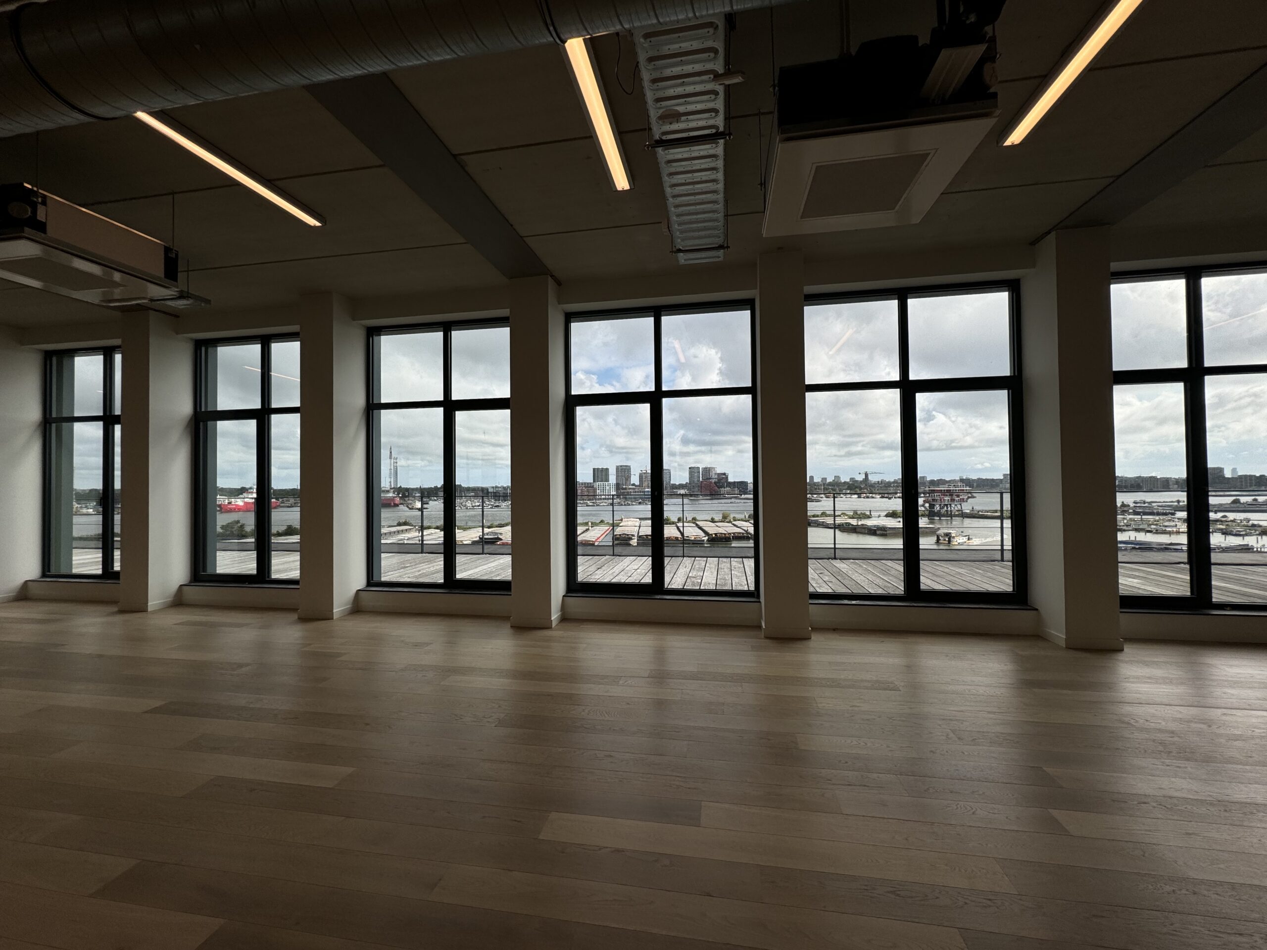 Een leeg interieur met grote ramen die uitkijken op een havengebied.
