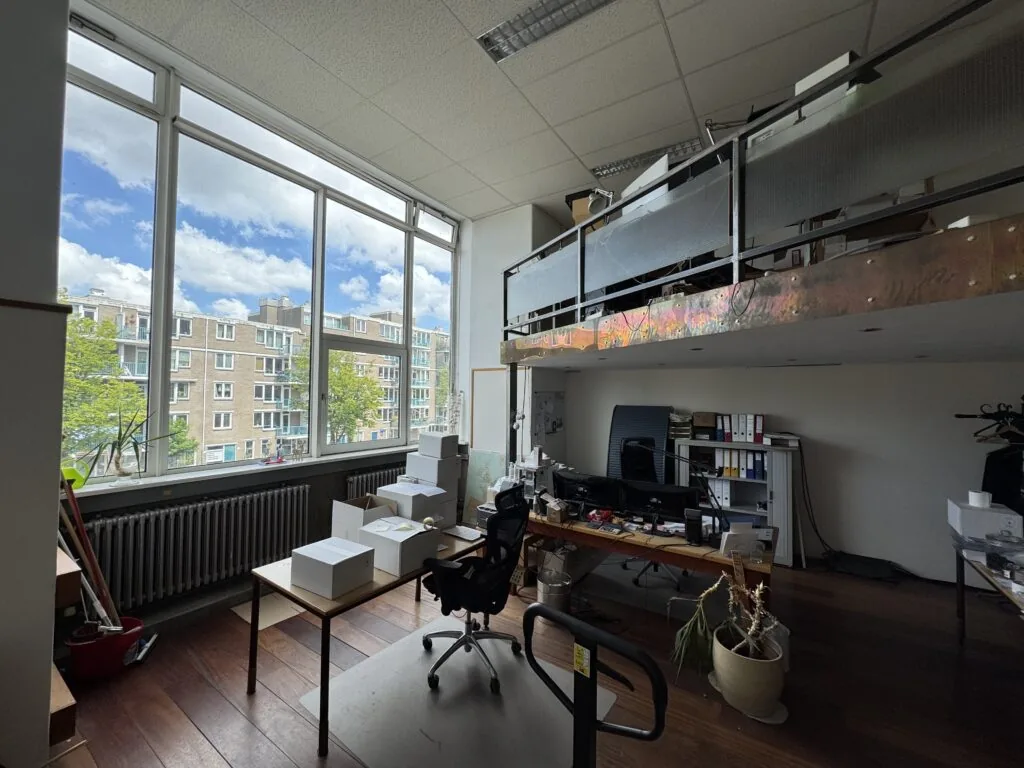 Een licht kantoor met grote ramen, bureaus, kartonnen dozen en een vide met opbergruimte.