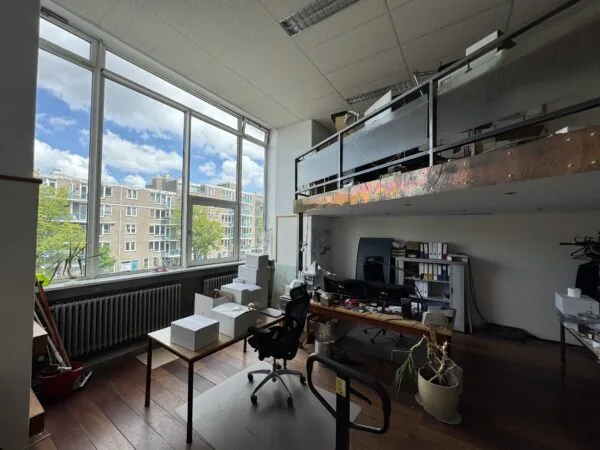 Een licht kantoor met grote ramen, bureaus, kartonnen dozen en een vide met opbergruimte.