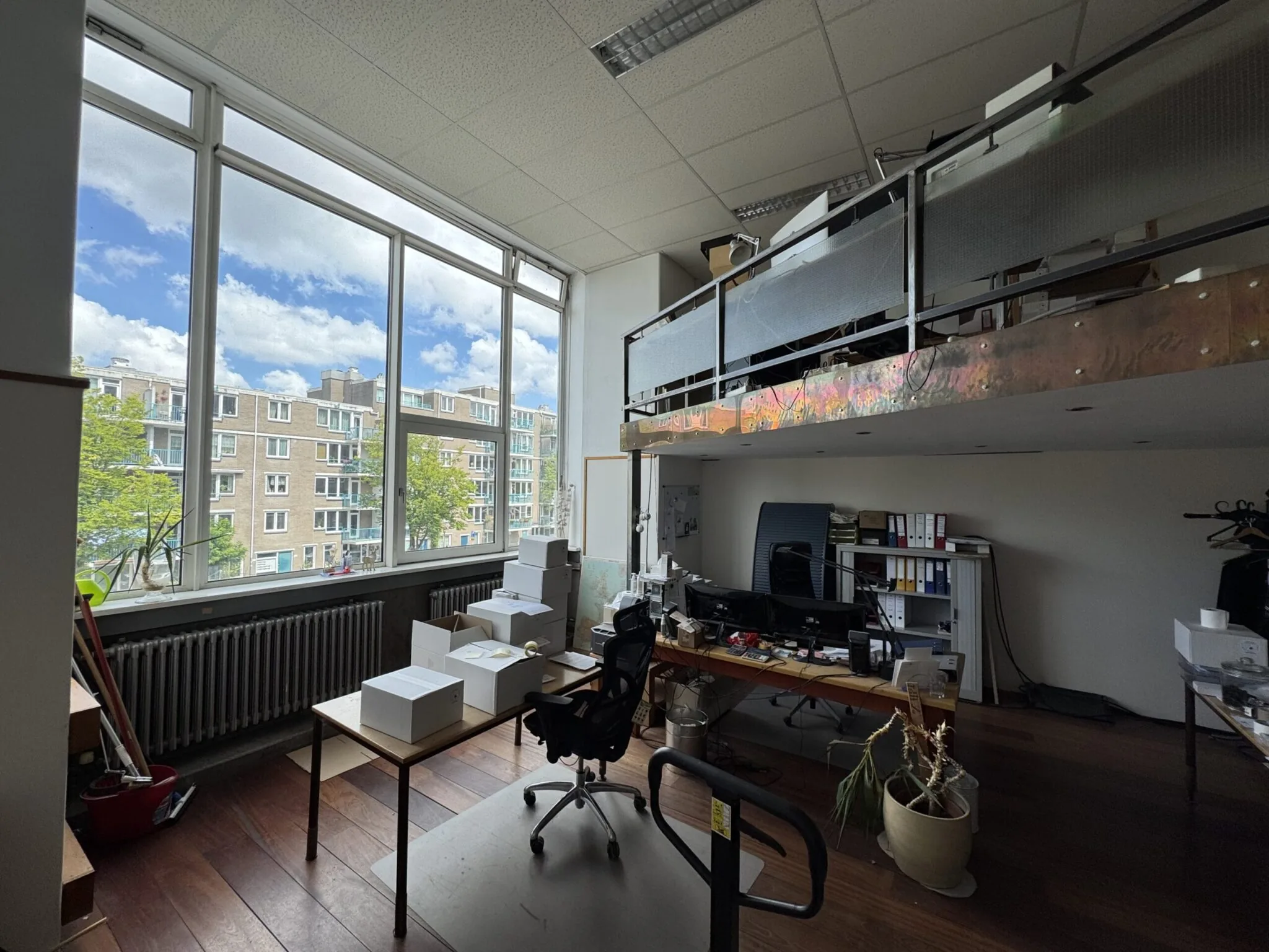 Een licht kantoor met grote ramen, bureaus, kartonnen dozen en een vide met opbergruimte.