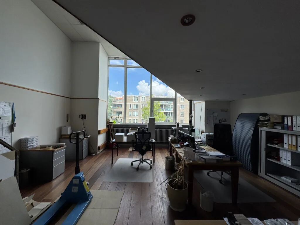 Een ruim en licht kantoor met houten vloer, bureaus, kantoorstoelen en een groot raam met uitzicht op een appartementencomplex.