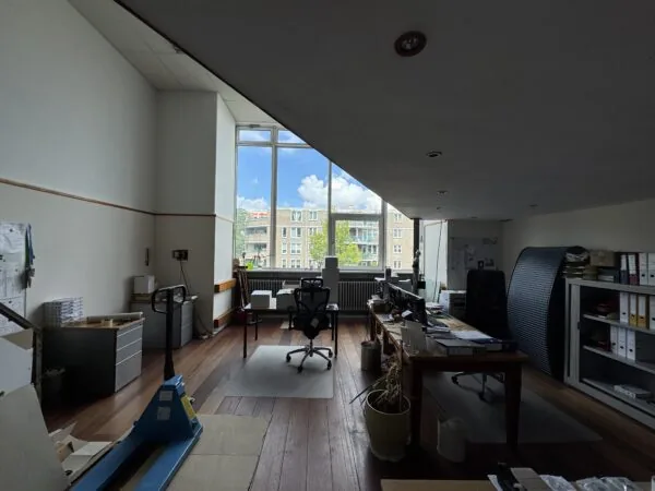 Een ruim en licht kantoor met houten vloer, bureaus, kantoorstoelen en een groot raam met uitzicht op een appartementencomplex.