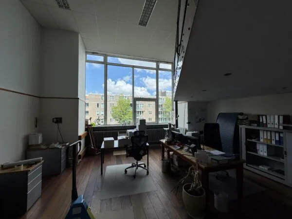 Een modern kantoor met houten vloer, bureaus en een groot raam met uitzicht op een woonwijk.