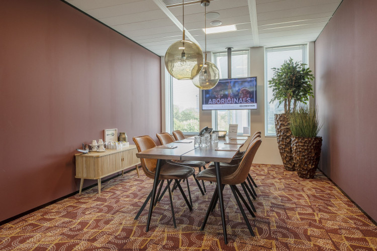 Vergaderruimte met houten tafel, lederen stoelen, grote planten en een tv-scherm aan de muur.