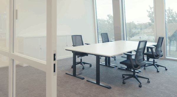 Vergaderruimte met een witte tafel, vijf zwarte bureaustoelen en grote ramen met uitzicht op bomen en een weg.