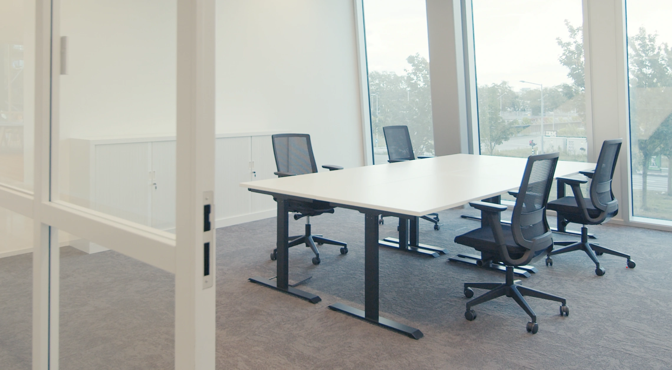 Vergaderruimte met een witte tafel, vijf zwarte bureaustoelen en grote ramen met uitzicht op bomen en een weg.