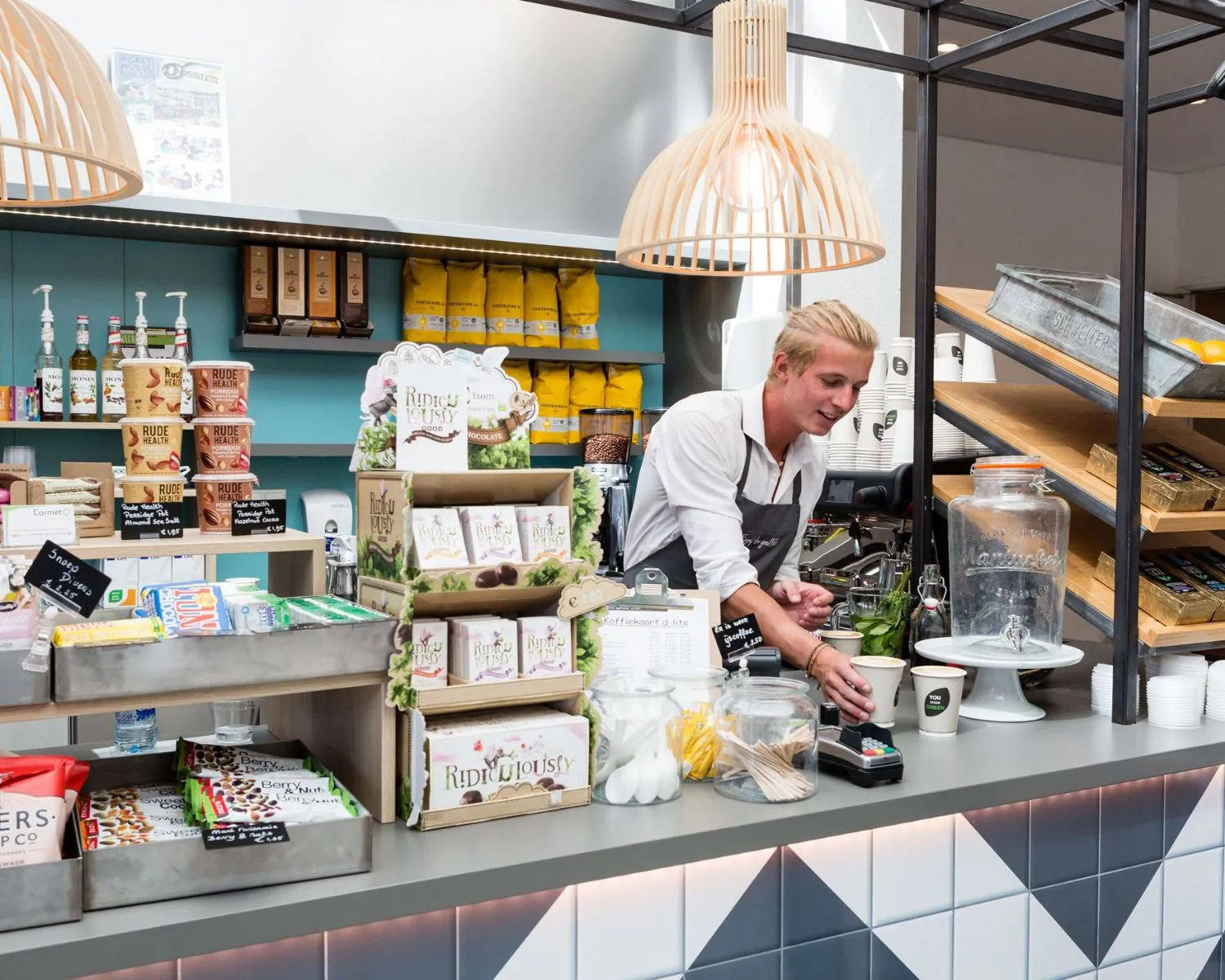 Man achter de koffiebar in een moderne bedrijfskantine aan de Capellalaan 65 in Hoofddorp, omringd door snacks en dranken.