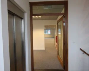 Ingang van een moderne kantoorruimte aan de De Ruijterkade 142 in Amsterdam met zicht op een lift en een glazen deur.