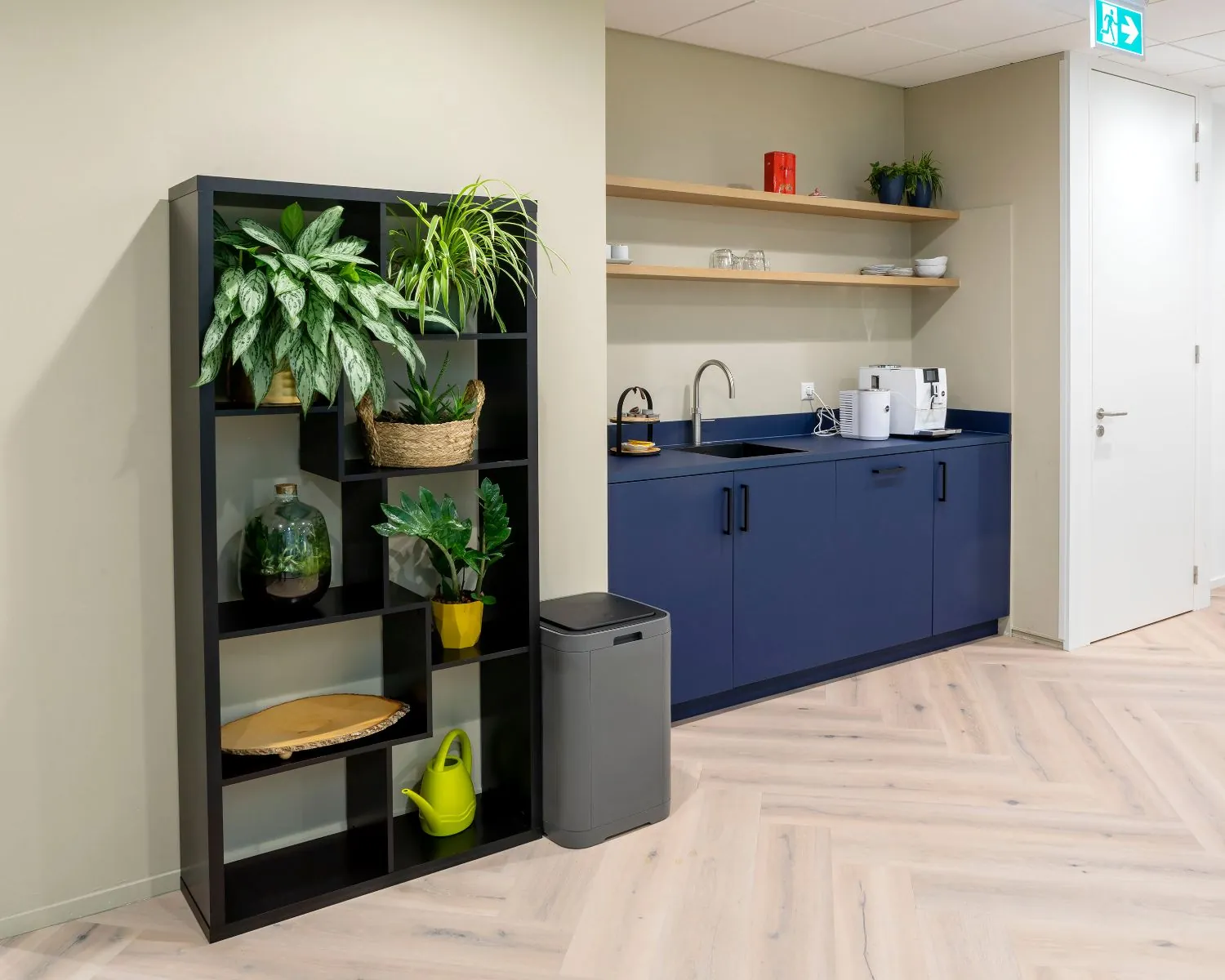 Moderne pantry in kantoorruimte aan de Kabelweg 57 in Amsterdam met blauwe keukenkastjes, open wandplanken en een zwarte plantenstelling.
