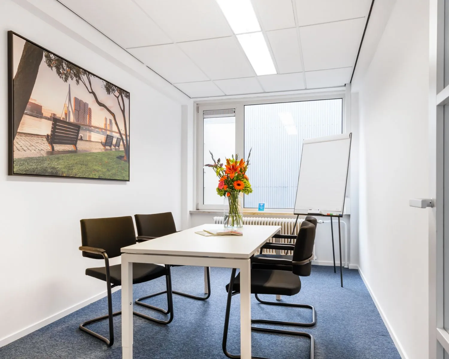Kleine vergaderruimte aan de Posthoornstraat 17 in Rotterdam met een tafel, vier stoelen, flip-over, bloemenvaas en een wandfoto van de Erasmusbrug.