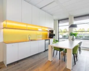 Moderne pantry met gele achterwand, witte kasten en een vergadertafel met groene stoelen in kantoorruimte aan de Databankweg 26 in Amersfoort.