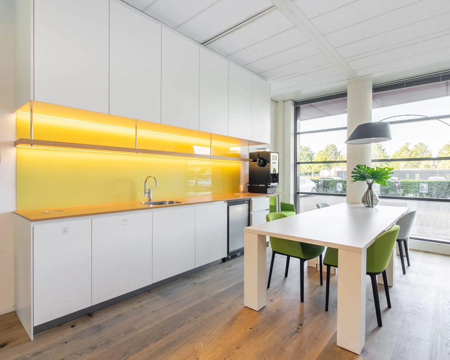 Moderne pantry met gele achterwand, witte kasten en een vergadertafel met groene stoelen in kantoorruimte aan de Databankweg 26 in Amersfoort.