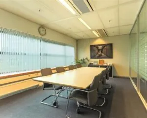 Vergaderruimte met lange tafel, acht stoelen, grote ramen met jaloezieën en een kunstwerk aan de muur in kantoor aan Avenue Céramique 221.