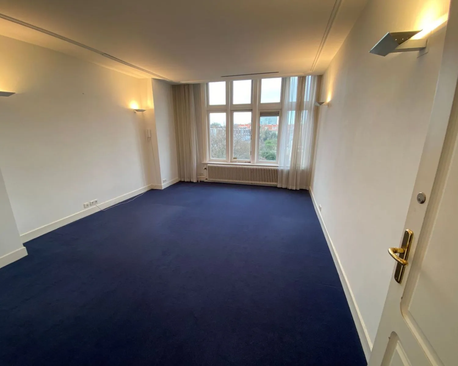 Lichte kantoorruimte te huur aan de Benoordenhoutseweg 21 met donkerblauwe vloerbedekking en grote ramen met uitzicht op de stad.