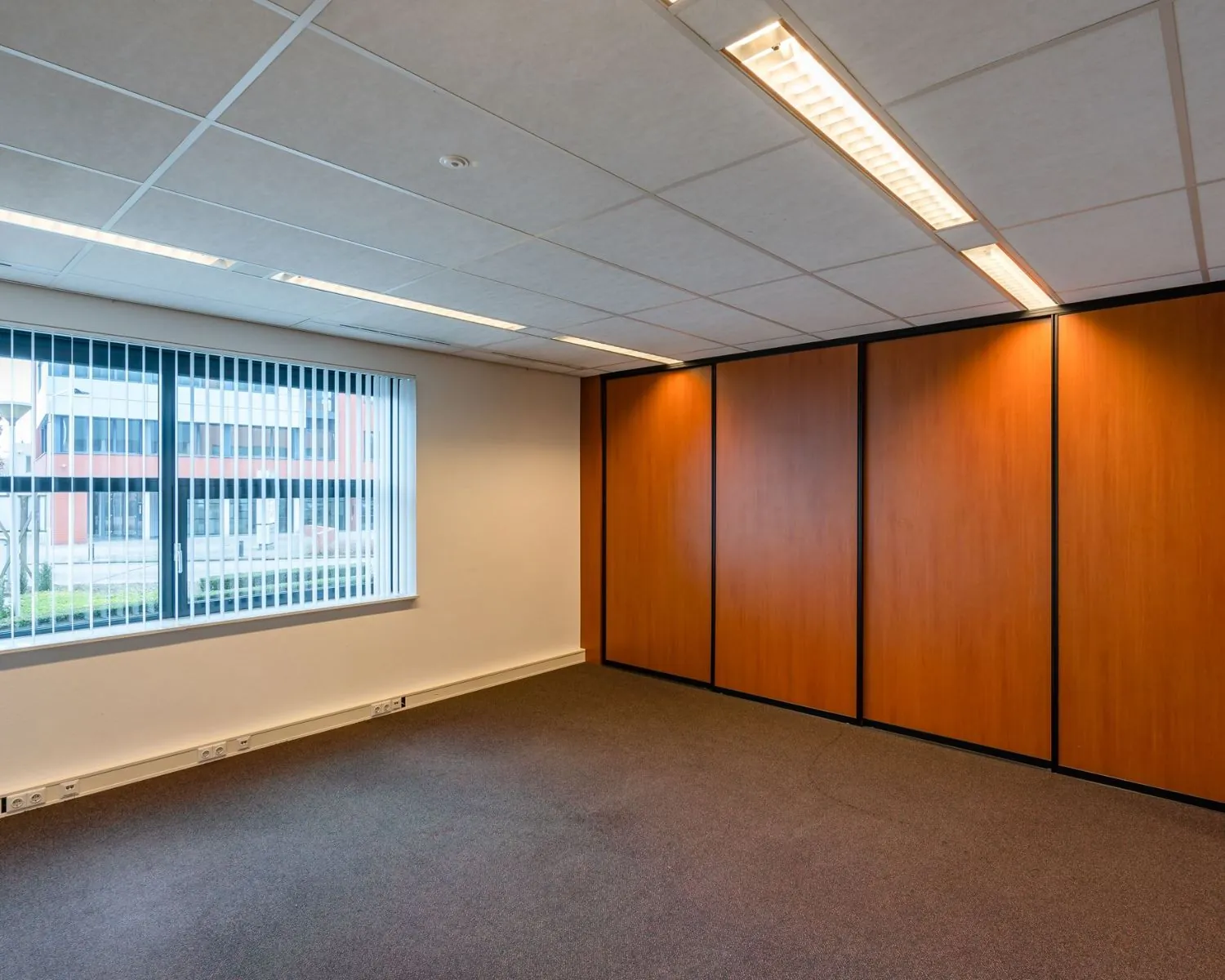Leegstaande kantoorruimte met tapijtvloer, systeemplafond en een groot raam met verticale jaloezieën in De Waal 18.