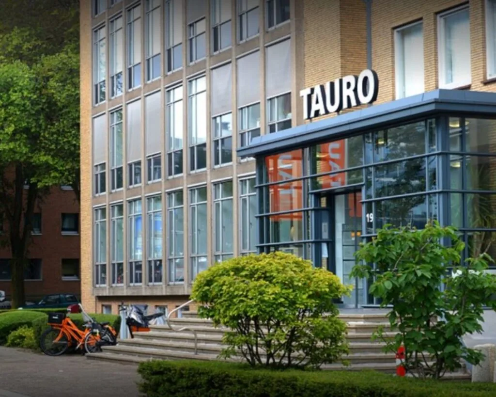 Vooraanzicht van het kantoorgebouw TAURO aan de President Kennedylaan 19 in Den Haag, met fietsen en groenvoorziening bij de entree.