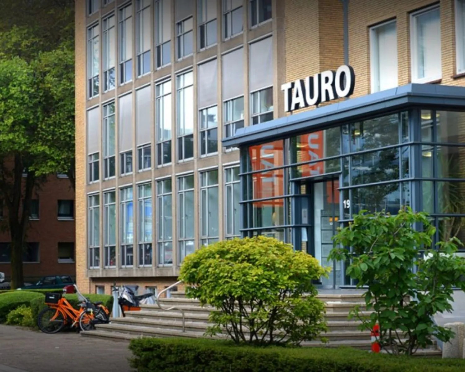 Vooraanzicht van het kantoorgebouw TAURO aan de President Kennedylaan 19 in Den Haag, met fietsen en groenvoorziening bij de entree.