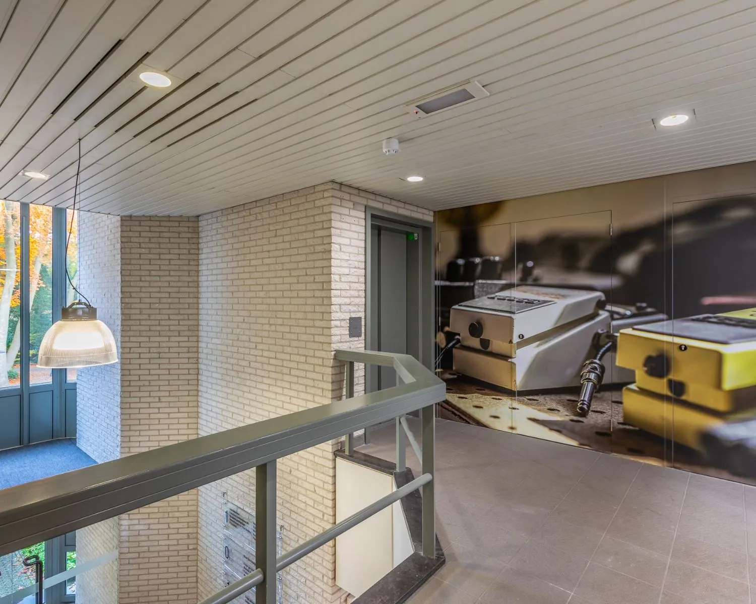 Moderne, lichte hal bij kantoorruimte aan de Mozartlaan 27 in Hilversum met lift en opvallende muurschildering van muziekapparatuur.