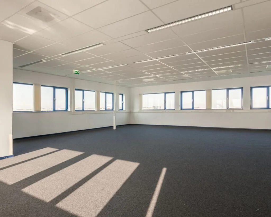 Lichte, lege kantoorruimte met blauw tapijt en grote ramen aan de Capronilaan in Schiphol-Rijk.